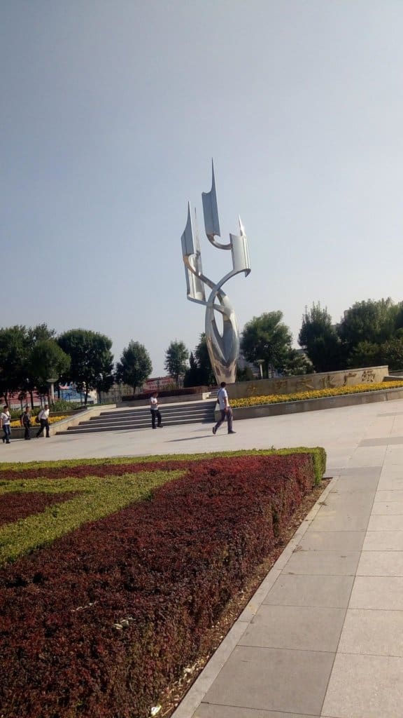 Crazy looking statue by the entrance to the subway near Shun Yi. People are walking along the path in front of the statue. The green and red grass is well kept along the paths and around the statue in a circular formation