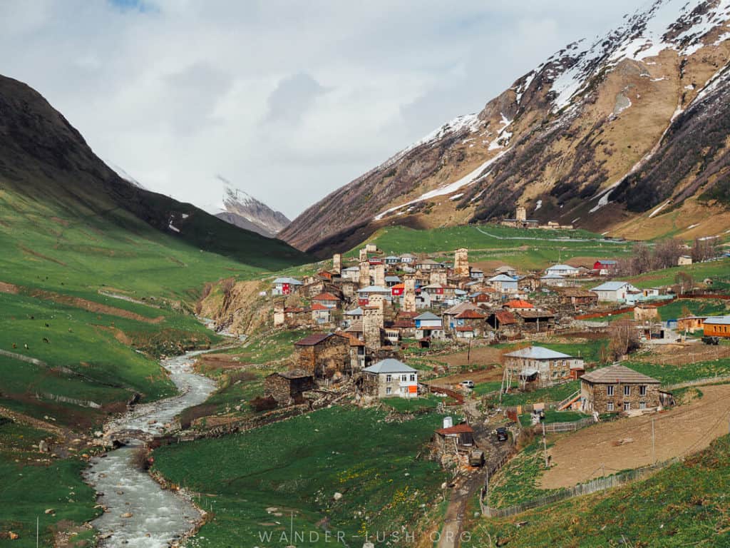 A picture of the small village of Ushguli, Georgia. Small stone houses make up a village surrounded bygreen grass and brown mountains. a river flows on the left side of the village. Georgia is among the Cheapest places to visit post-pandemic