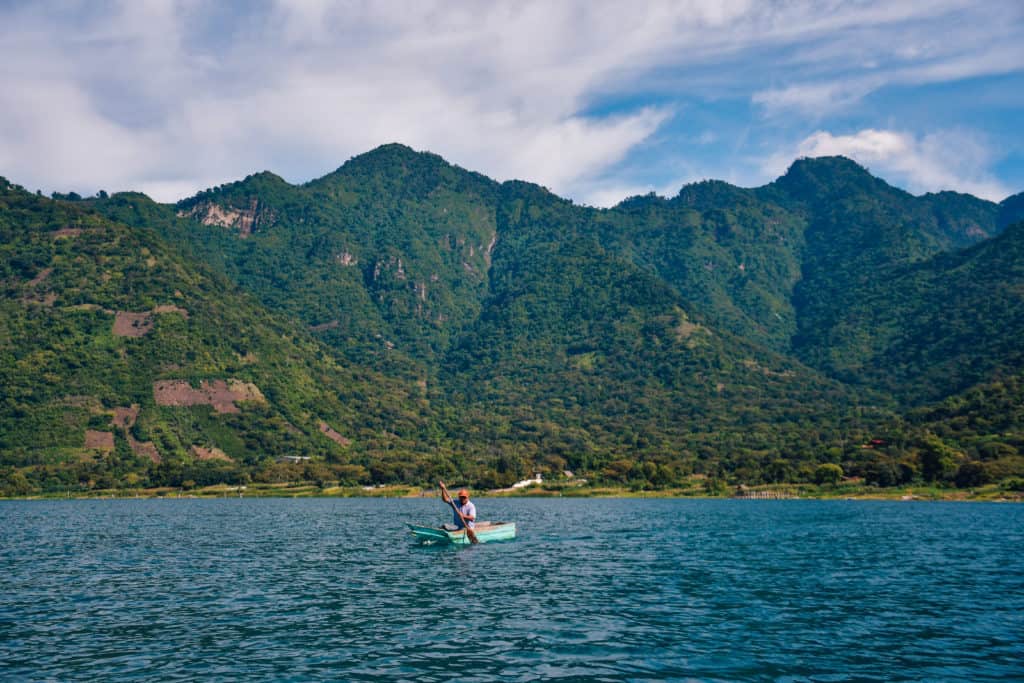 A man paddles a blue boat on water with towering green mountains in the background.
