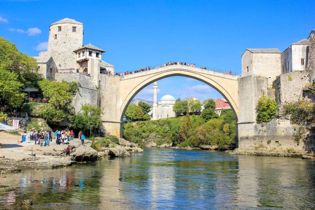 A small river passing under an old stone bridge with stone towers on either side. Under the bridge more stone buildings can be seen behind some trees and bushes. Mostar is one of the cheapest places to visit post-pandemic