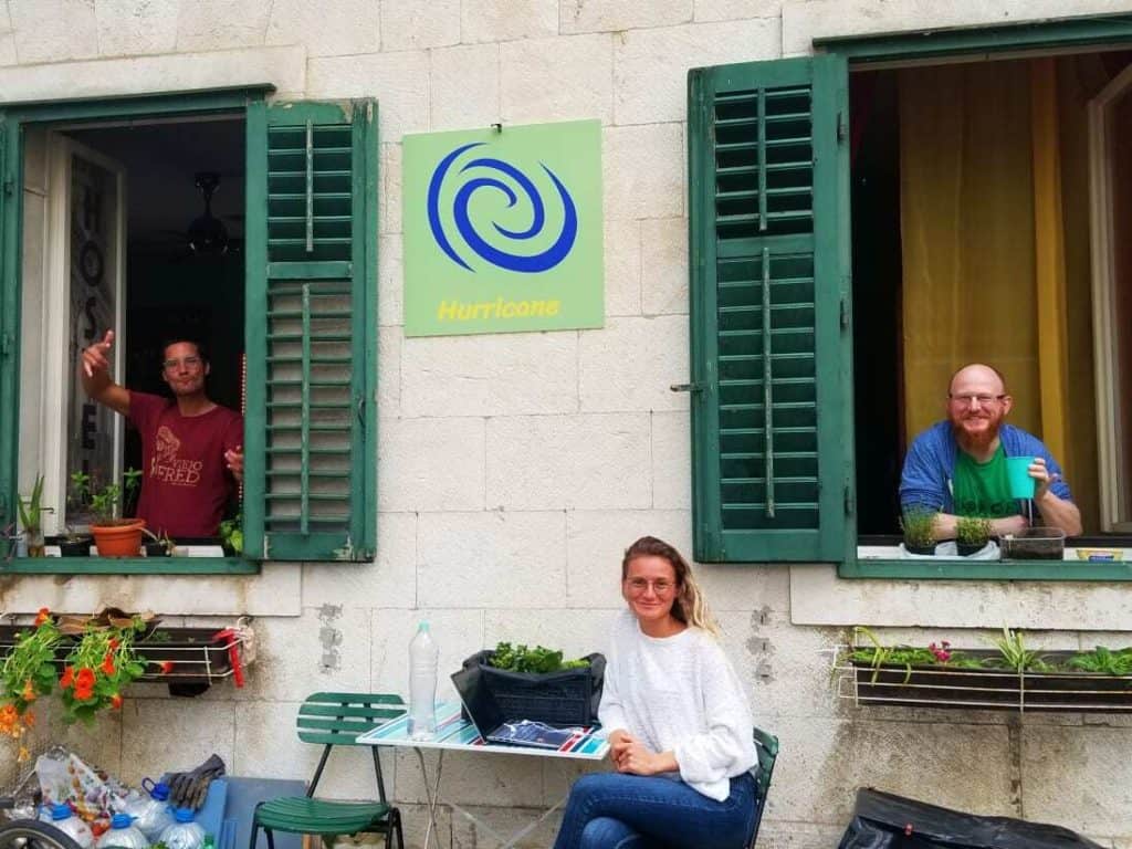 One person sitting out front of hurricane hostel underneath its sign. Two people are standing in the window on either side. one of the best places to stay around Croatia